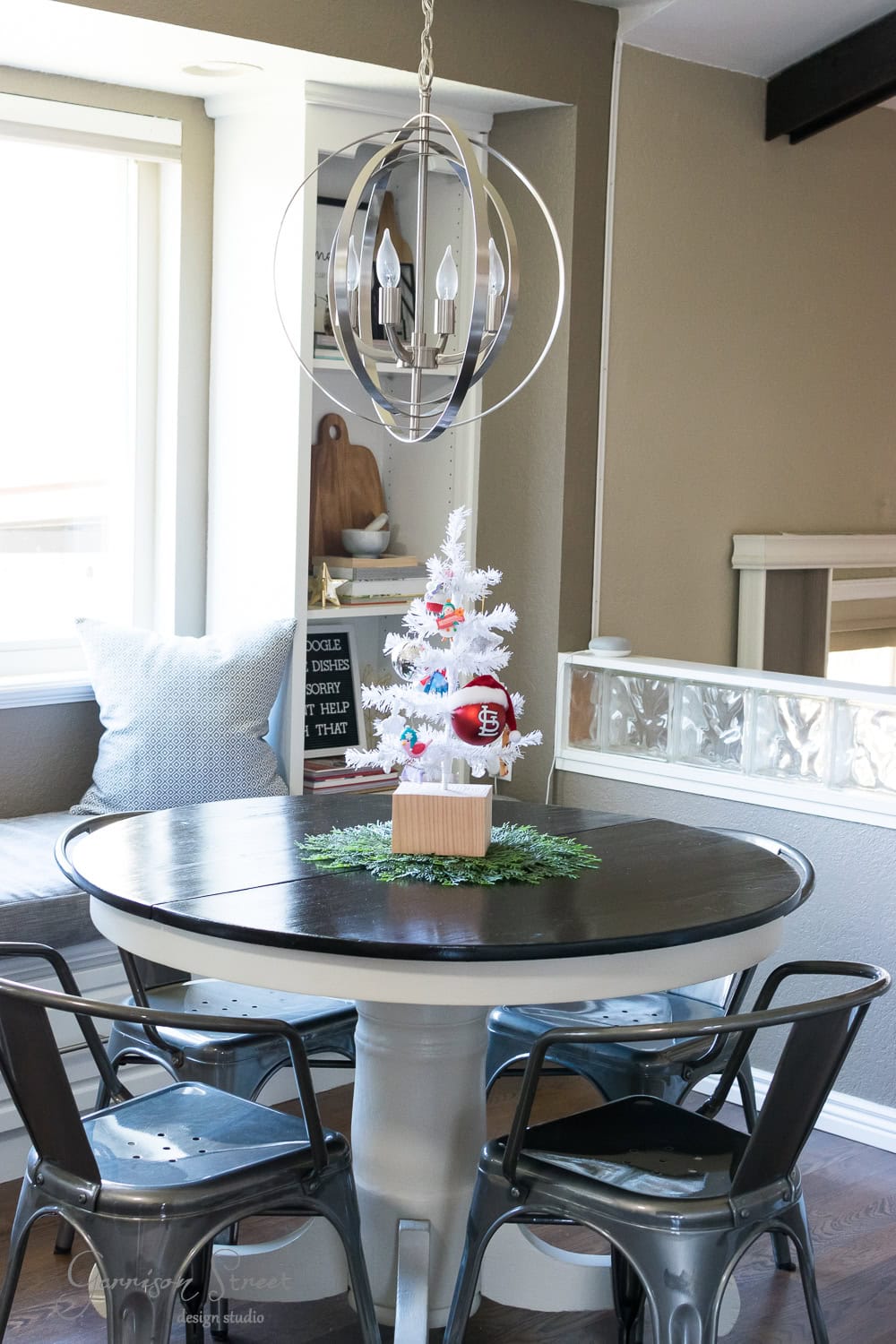 A cozy dining nook features a modern round black table with metal chairs. A small white Christmas tree with red ornaments sits on the table. Soft gray pillows adorn the built-in bench. A sleek chandelier hangs above, illuminating the space with a festive glow.