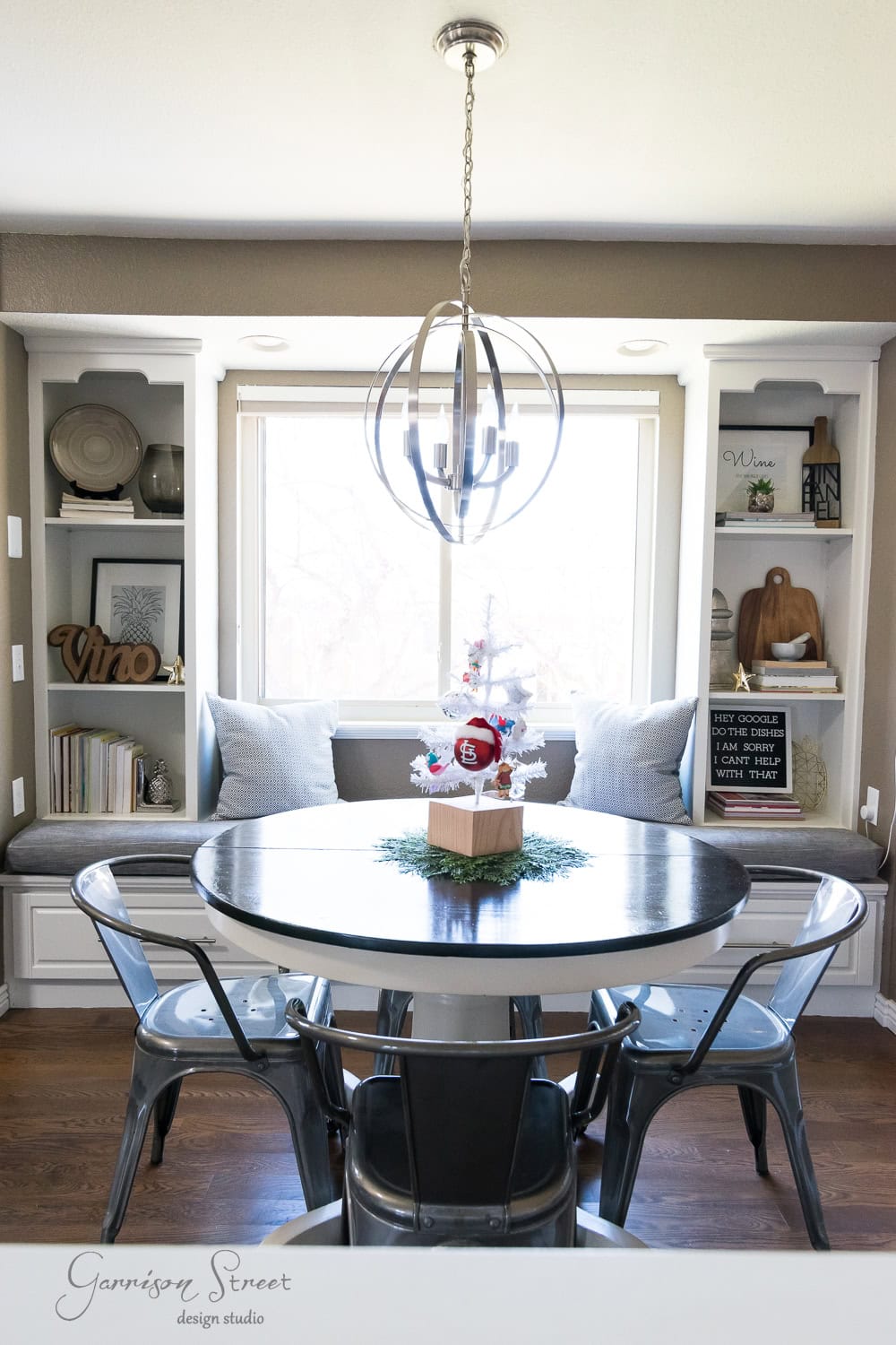 A cozy dining nook with a circular table surrounded by metal chairs and built-in shelving. A decorative Christmas centerpiece adorns the table, while a modern chandelier hangs above. Neutral tones and natural light enhance the inviting atmosphere.