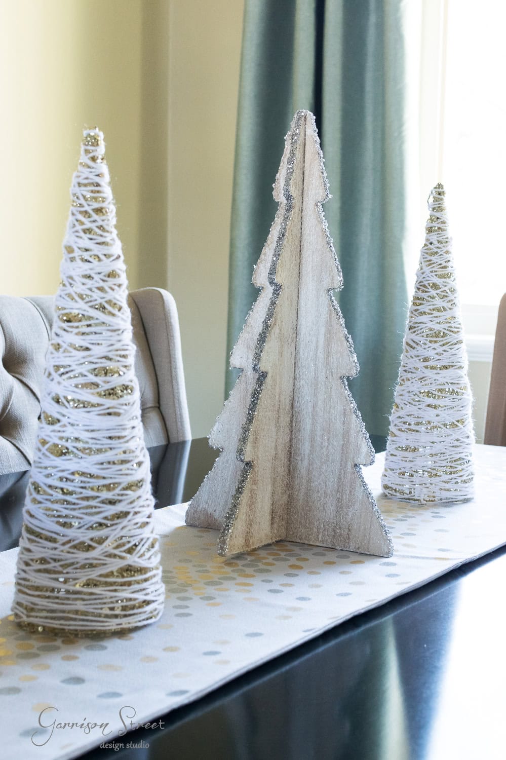 A modern table centerpiece features three Christmas trees: a wooden one in the middle and two wrapped in white yarn on each side. They rest on a dotted runner over the dark table, with a chair and curtained window subtly framing the festive scene.