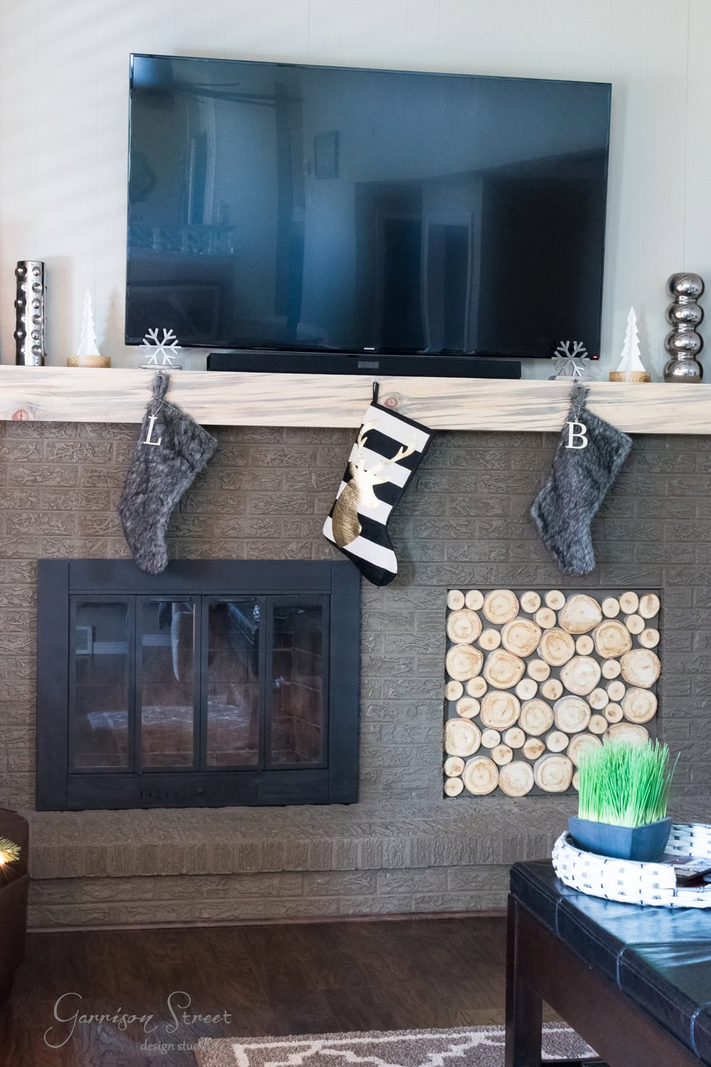 A modern living room features a TV mounted above a fireplace decorated with festive Christmas stockings and candles. The mantel showcases three stockings, with the letters "L" and "B" visible. Below, wood logs are arranged in a tidy tiled pattern, adding warmth to the sleek design.