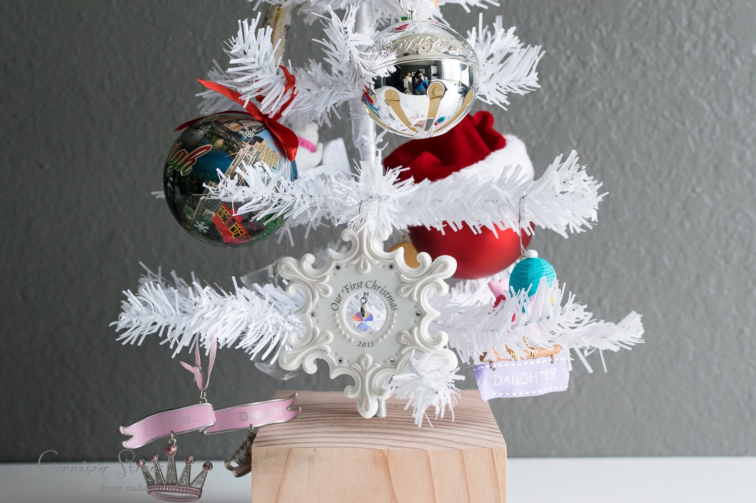 DIY Dollar Store Christmas Tree Hack A small white artificial Christmas tree with various ornaments, including a "First Christmas 2019" frame, a Santa hat, and a "Daughter" bauble. The tree is placed on a wooden stand against a gray background.