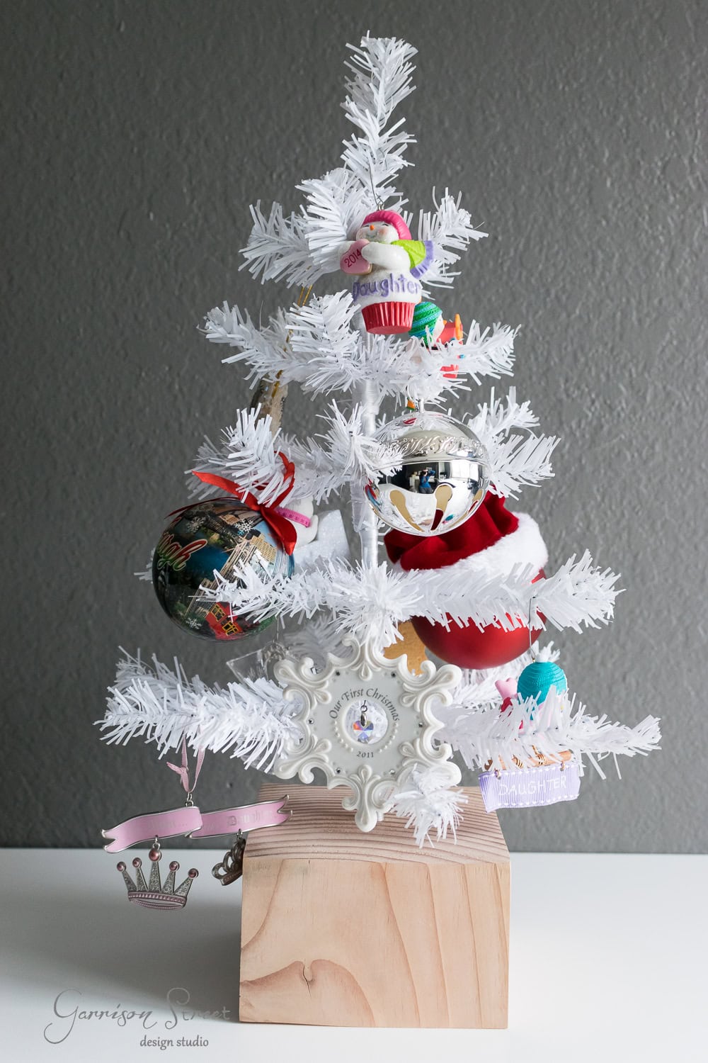 DIY Dollar Store Christmas Tree Hack A small white artificial Christmas tree stands on a wooden block. It is adorned with colorful ornaments, including a globe, a silver bauble, and Santas. A decorative frame with text is at the base. The background is a gray wall.