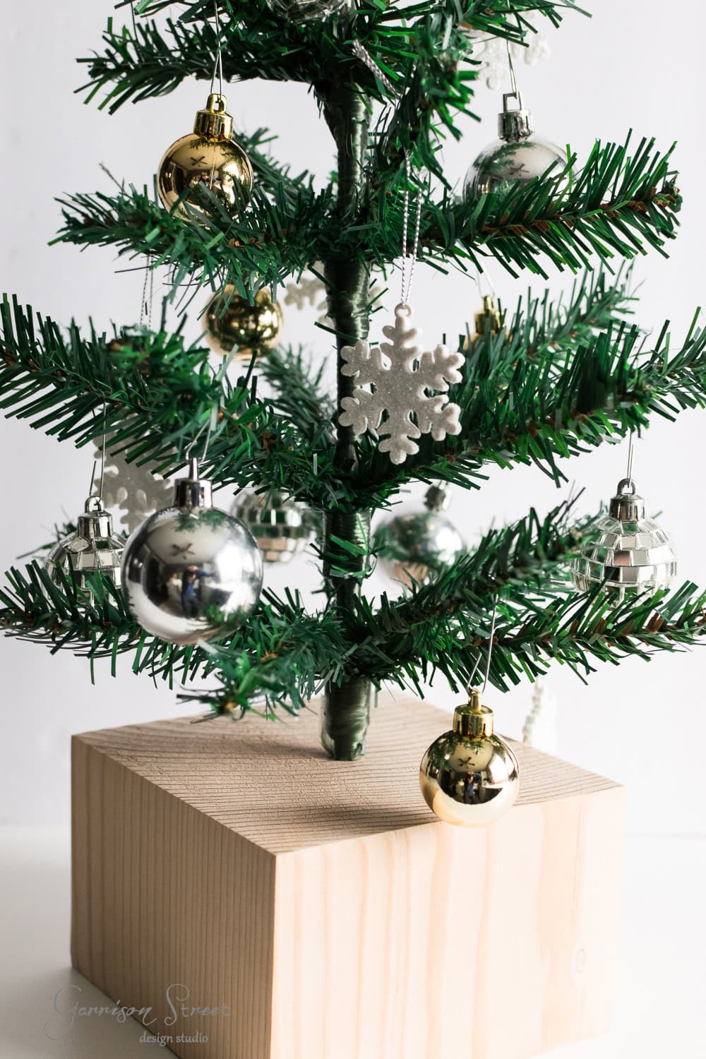 DIY Dollar Store Christmas Tree Hack A small Christmas tree with silver and gold ornaments, including a snowflake decoration, stands on a wooden block. The tree is set against a white background.