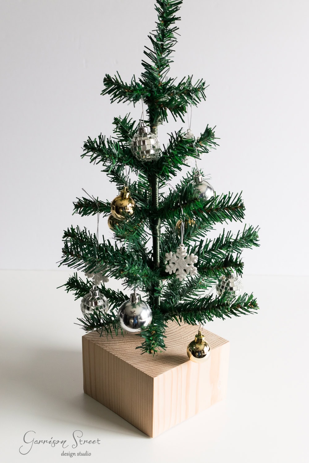 DIY Dollar Store Christmas Tree Hack A small artificial Christmas tree with silver and gold ornaments is placed on a wooden block. The ornaments include a snowflake and spherical decorative balls, creating a festive look against a plain background.