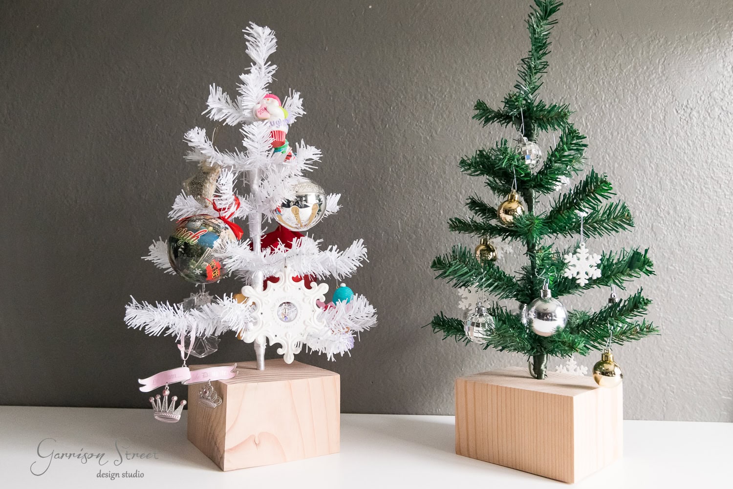 Two small Christmas trees, one white and one green, are decorated with various ornaments including baubles and snowflakes. They are set on wooden block stands against a neutral wall, with a signature reading "Garrison Street design studio.