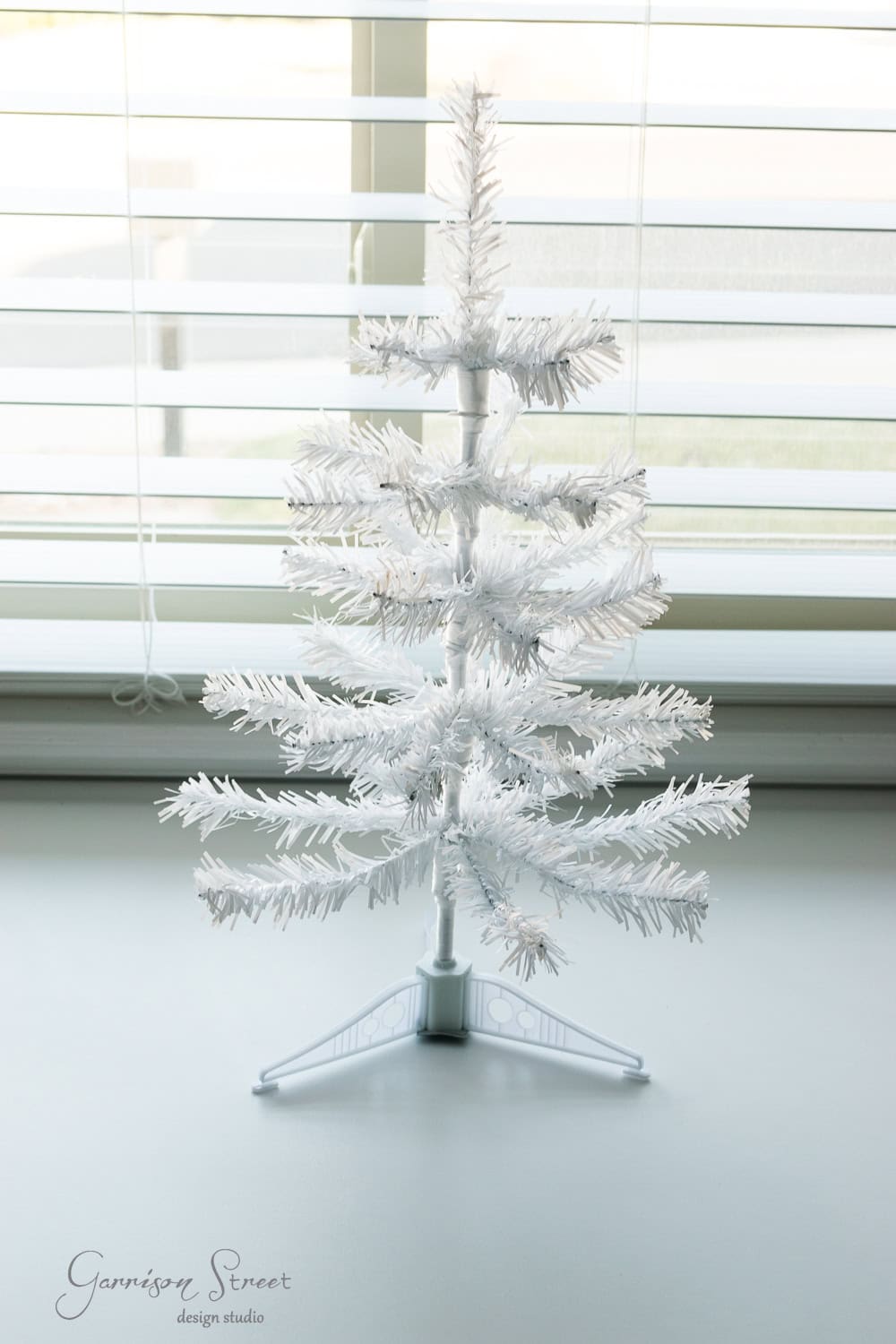 DIY Dollar Store Christmas Tree Hack A small, white artificial Christmas tree with sparse branches stands on a white surface in front of a window with blinds. Sunlight filters through, casting soft light on the tree.