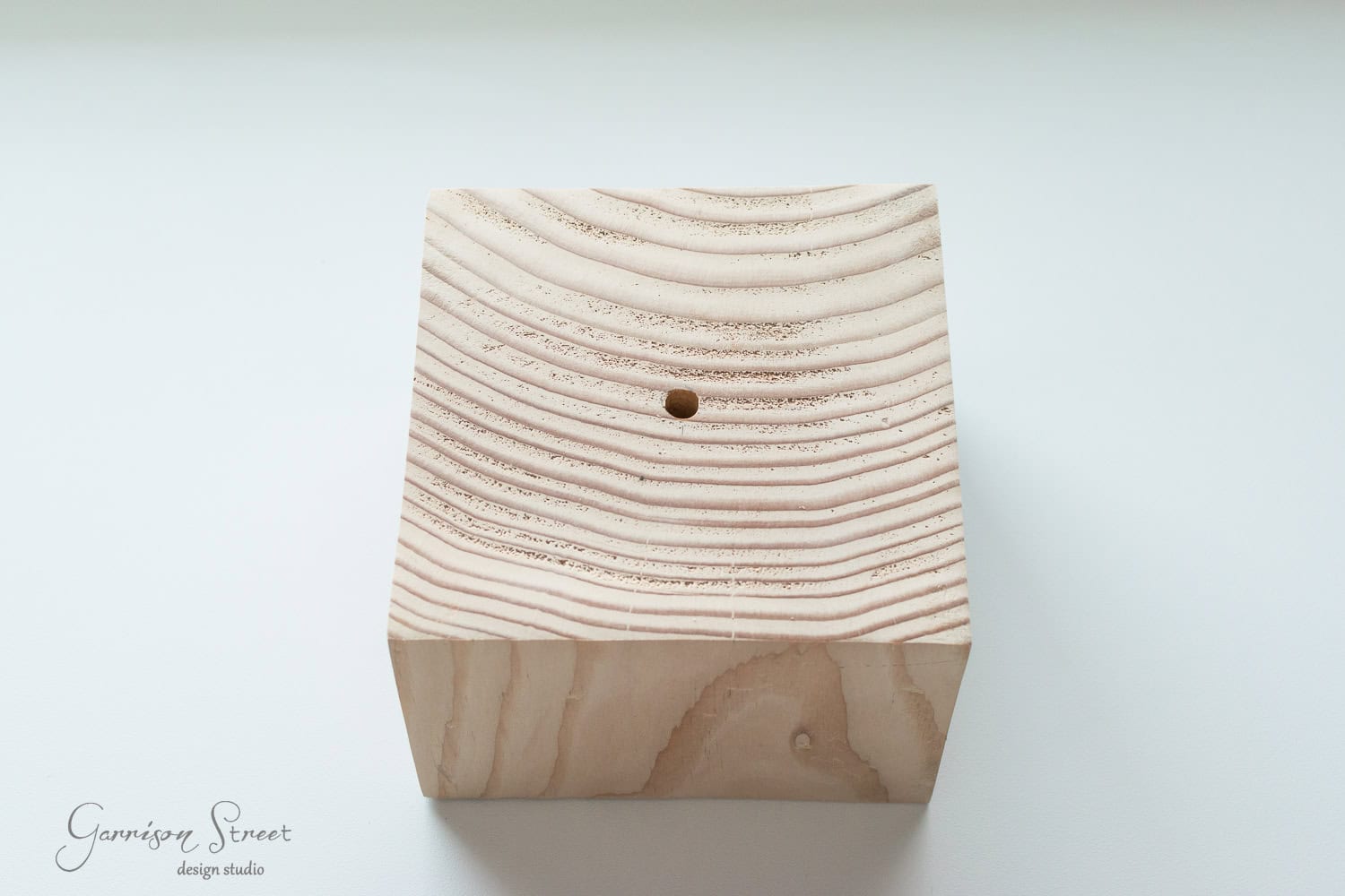 DIY Dollar Store Christmas Tree Hack A cube of light-colored wood with visible grain patterns and a small hole in the center, resting on a white surface. The logo "Garrison Street Design Studio" is in the bottom left corner.