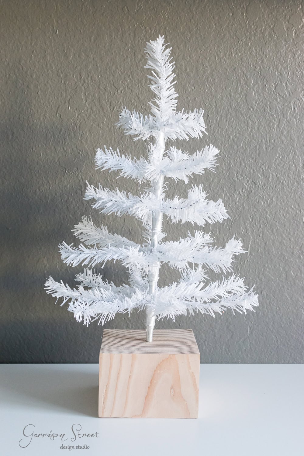DIY Dollar Store Christmas Tree Hack A small white artificial Christmas tree with sparse branches is mounted on a wooden block against a plain gray background.