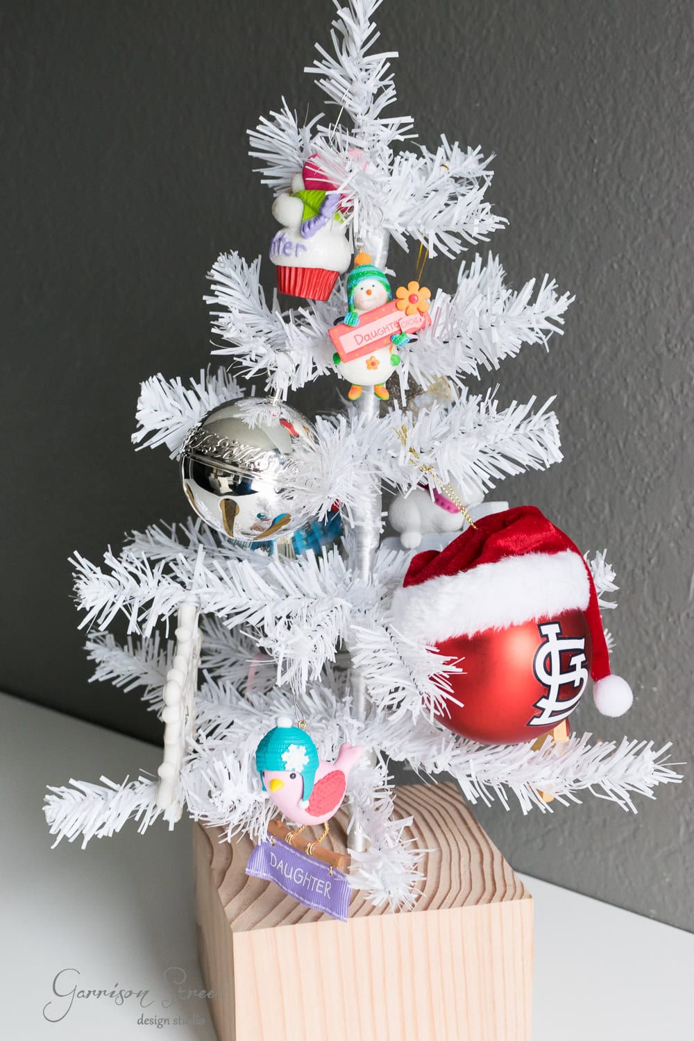 DIY Dollar Store Christmas Tree Hack A small white Christmas tree with various ornaments, including a red and white hat, a silver bauble, and items labeled "Daughter." The tree sits on a wooden block against a gray background.