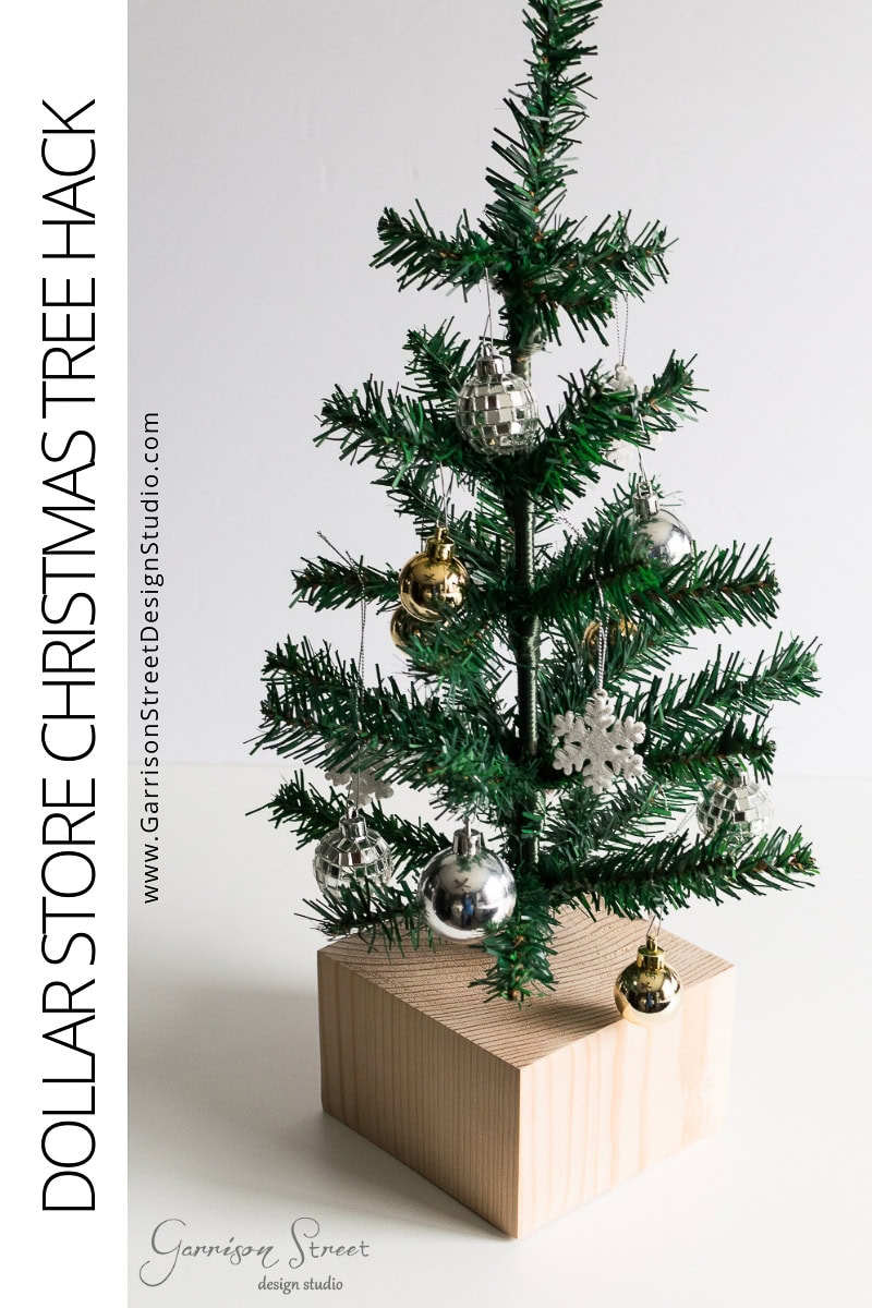 DIY Dollar Store Christmas Tree Hack A small artificial Christmas tree, decorated with silver and gold ornaments, sits elegantly on a wooden base. This easy decor idea is titled "Dollar Store Christmas Tree Hack" and includes a website link on the left for more details.