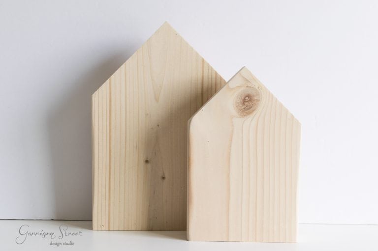 Two wooden blocks shaped like simple houses, one larger and one smaller, stand against a plain white background. The wood grain and knots are visible, adding texture to the minimalistic design.