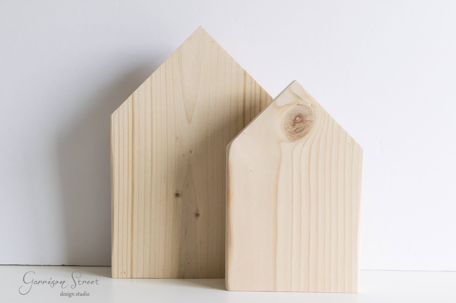 Two wooden blocks shaped like simple houses, one larger and one smaller, stand against a plain white background. The wood grain and knots are visible, adding texture to the minimalistic design.