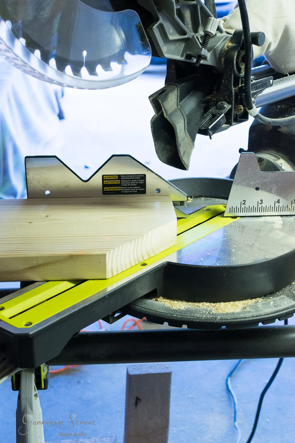 A power miter saw is cutting a piece of wood on a workbench. Safety warnings are visible on the saw, and there is a ruler for precision measurement. The workspace has scattered sawdust.