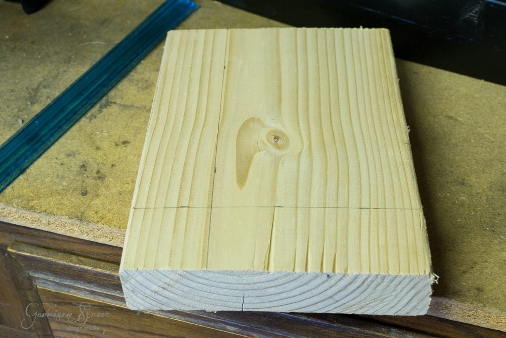 A wooden plank with a natural knot sits on a workbench. Two pencil lines are drawn across its surface, and a blue measuring tool is partially visible beside it.