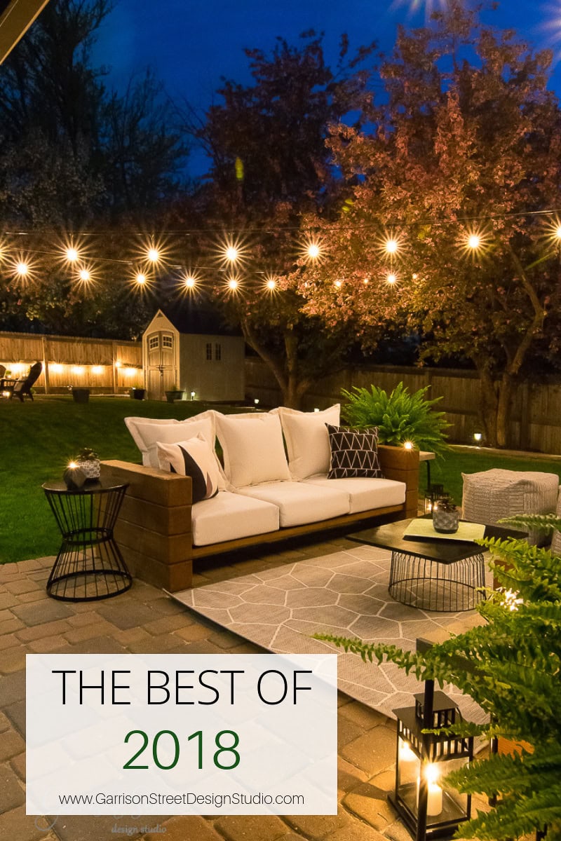A cozy outdoor patio at dusk with string lights, a cushioned sofa, and stylish tables. Lush greenery surrounds, with a small lit structure in the background. Text reads "The Best of 2018" from Garrison Street Design Studio.