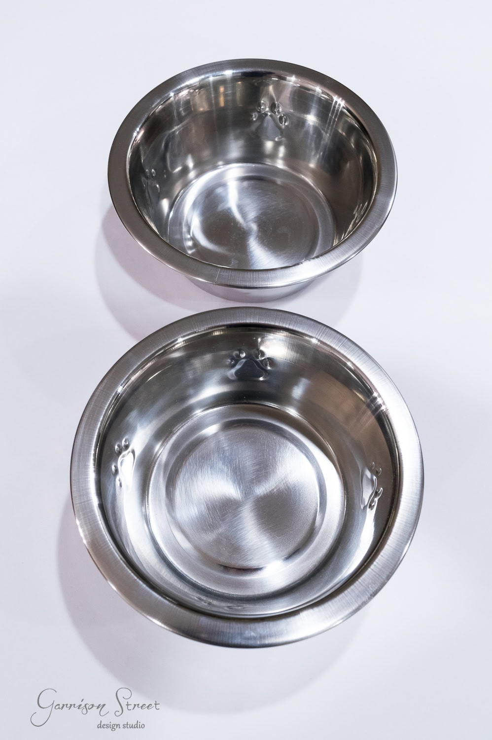 Two stainless steel dog bowls are placed on a white surface. They are shiny and reflective, with a simple, clean design. The raised edges are visible as they slightly nest together, one partially overlapping the other.