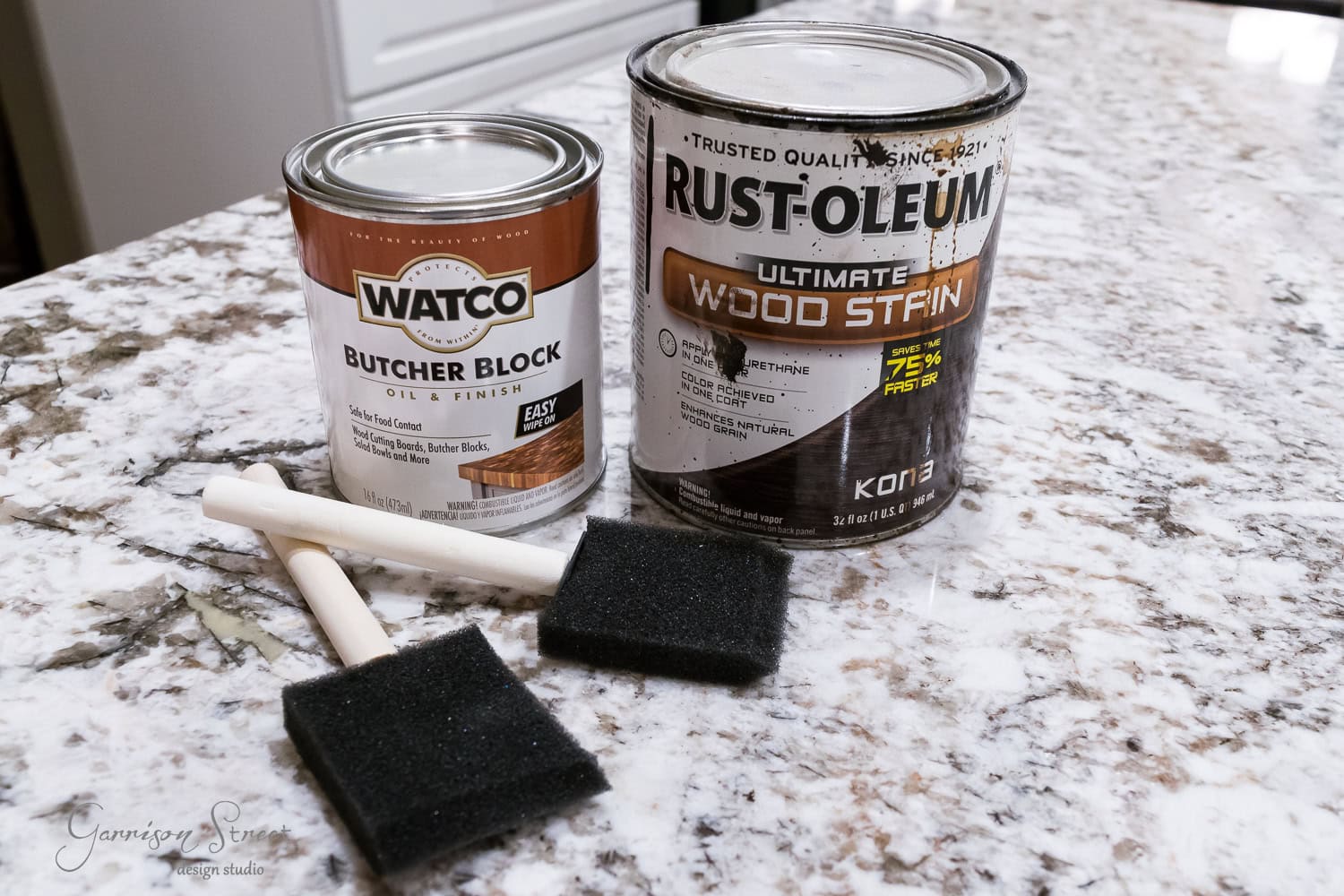 Two cans of finish rest on a marble countertop: Watco Butcher Block oil finish and Rust-Oleum wood stain. In front, two foam brushes await, ready to bring new life to a raised dog bowl or any wood surface needing care.