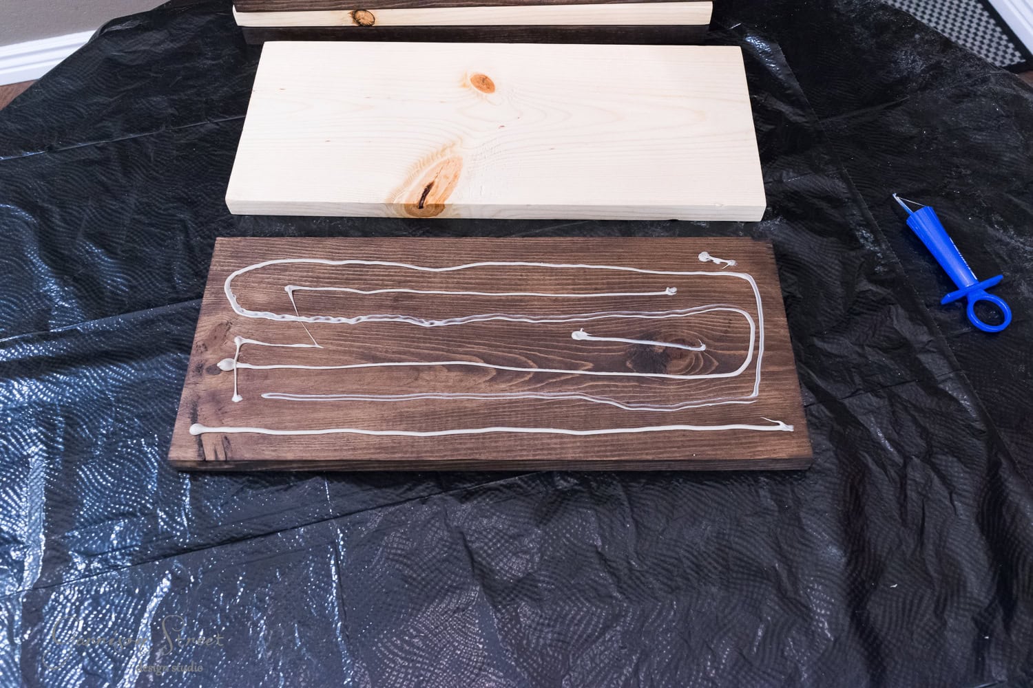 Two wooden boards on a black plastic sheet: one plain, one stained dark brown with zigzagging glue lines. A blue knife with a safety cover lies beside them.