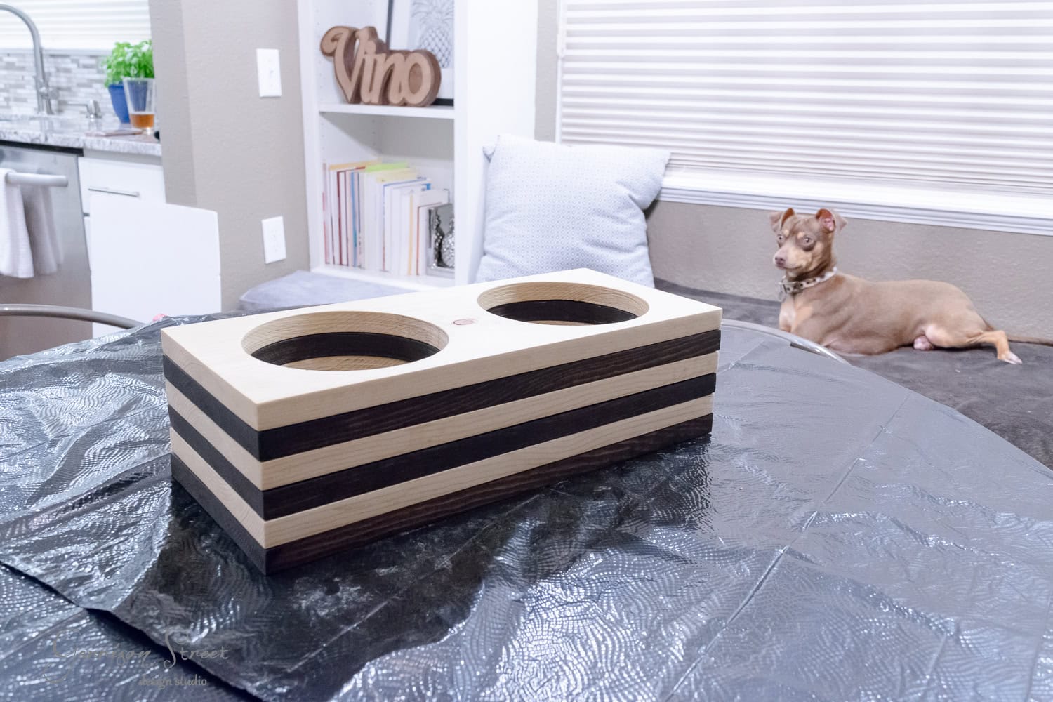 A wooden raised pet bowl holder with two circular openings sits on a covered table. A small dog rests in the background on a cushioned chair. The room features a bookshelf, a "Vino" sign, and a partially visible kitchen area.