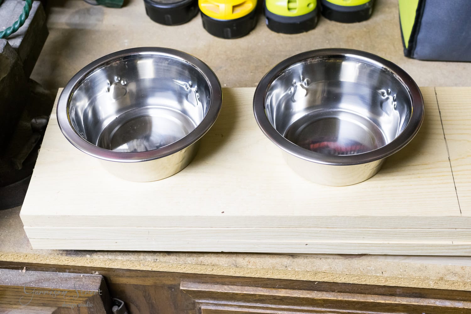 Two shiny, raised stainless steel dog bowls sit side by side on a wooden board. The setup is on a workbench, with tools and equipment visible in the background, suggesting a workshop environment.