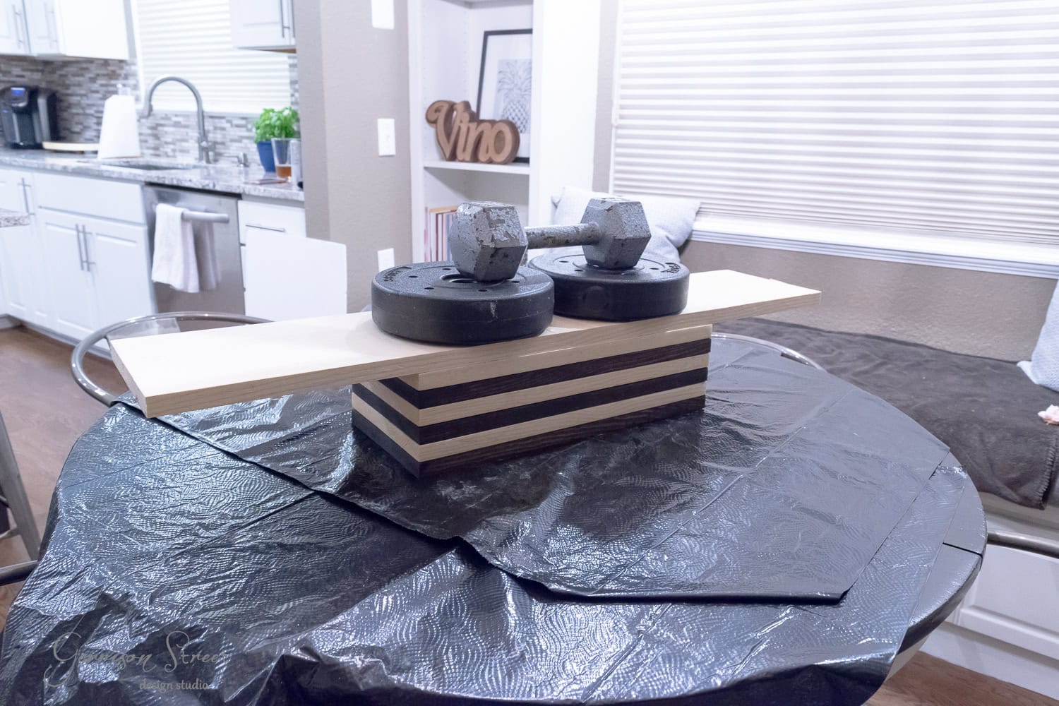 A DIY clamping setup features a wooden board balanced on small fitness weights and a dumbbell in a kitchen. The scene includes a raised, rounded table covered with a black cloth, and there's even a dog bowl peeking from the background.