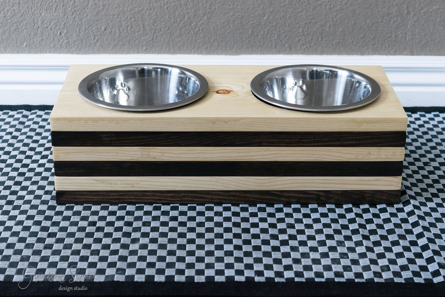 Two stainless steel dog bowls are set in a raised wooden stand with alternating light and dark stripes, resting on a black and white checkered mat against a neutral wall.