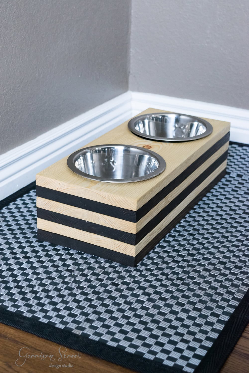 A wooden elevated pet feeder for your dog, featuring two stainless steel bowls, sits on a patterned black and white mat in a corner. The feeder's striped design complements the light gray wall with white trim.