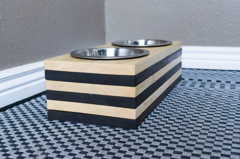 A stylish raised pet feeder with two metal bowls, perfect for your dog, set on a geometric-patterned mat. The feeder boasts alternating light wood and dark stripes, neatly placed in the corner against a gray wall.