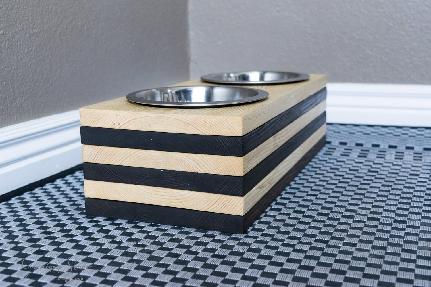 A stylish raised pet feeder with two metal bowls, perfect for your dog, set on a geometric-patterned mat. The feeder boasts alternating light wood and dark stripes, neatly placed in the corner against a gray wall.