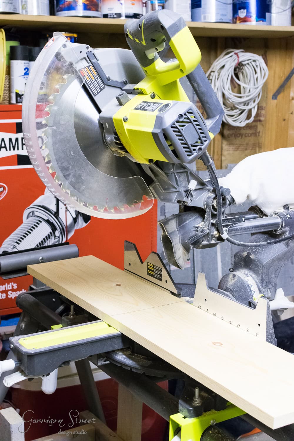 A green and silver miter saw sits on a raised workbench with a wooden board placed on it. In the background, there are shelves with paint cans and coiled cables. The workspace appears organized, tools neatly arranged.