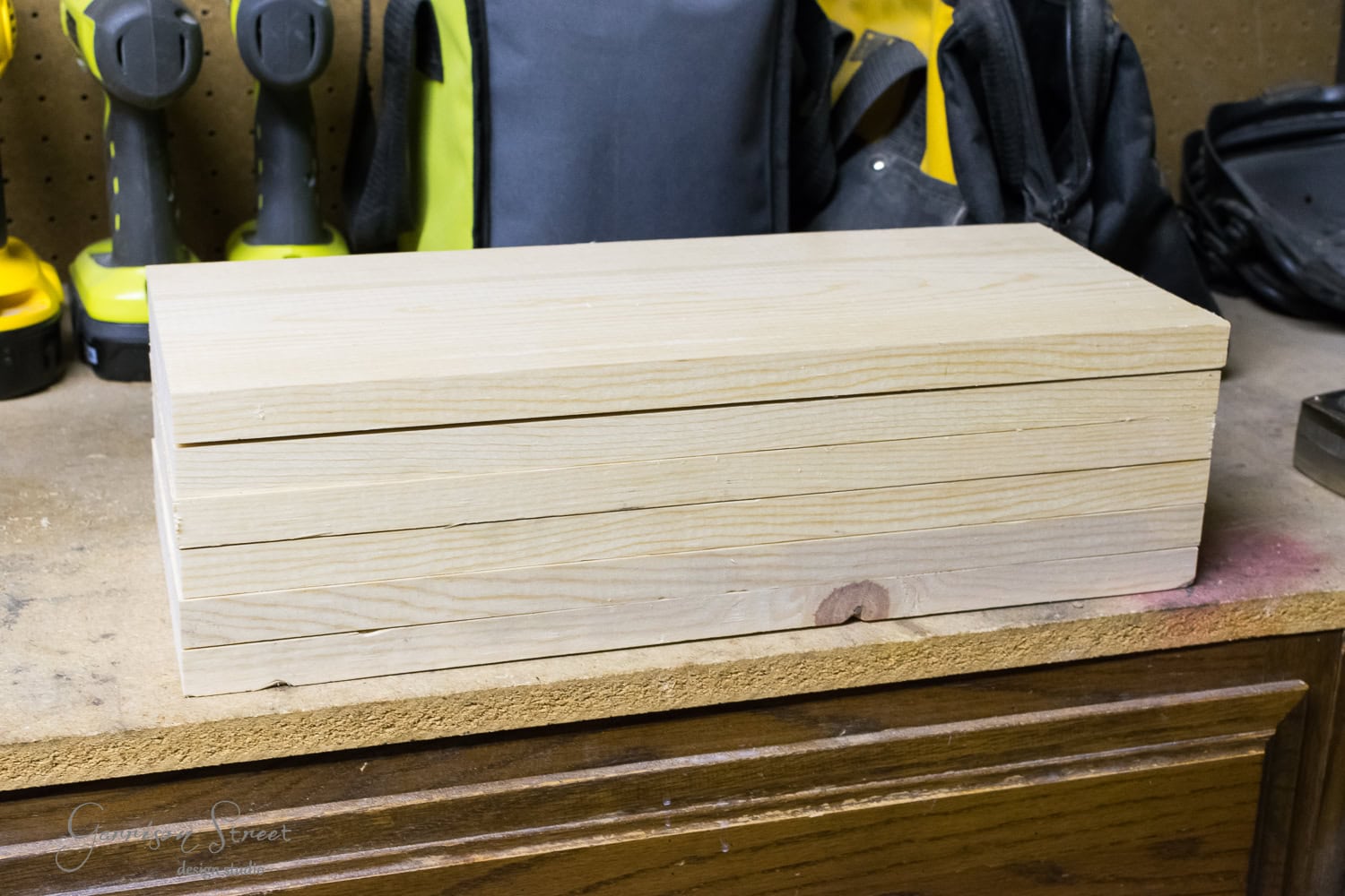A stack of three wooden boards rests on the workbench, surrounded by tools like drills and bags. The unfinished boards are neatly placed on the wooden surface.