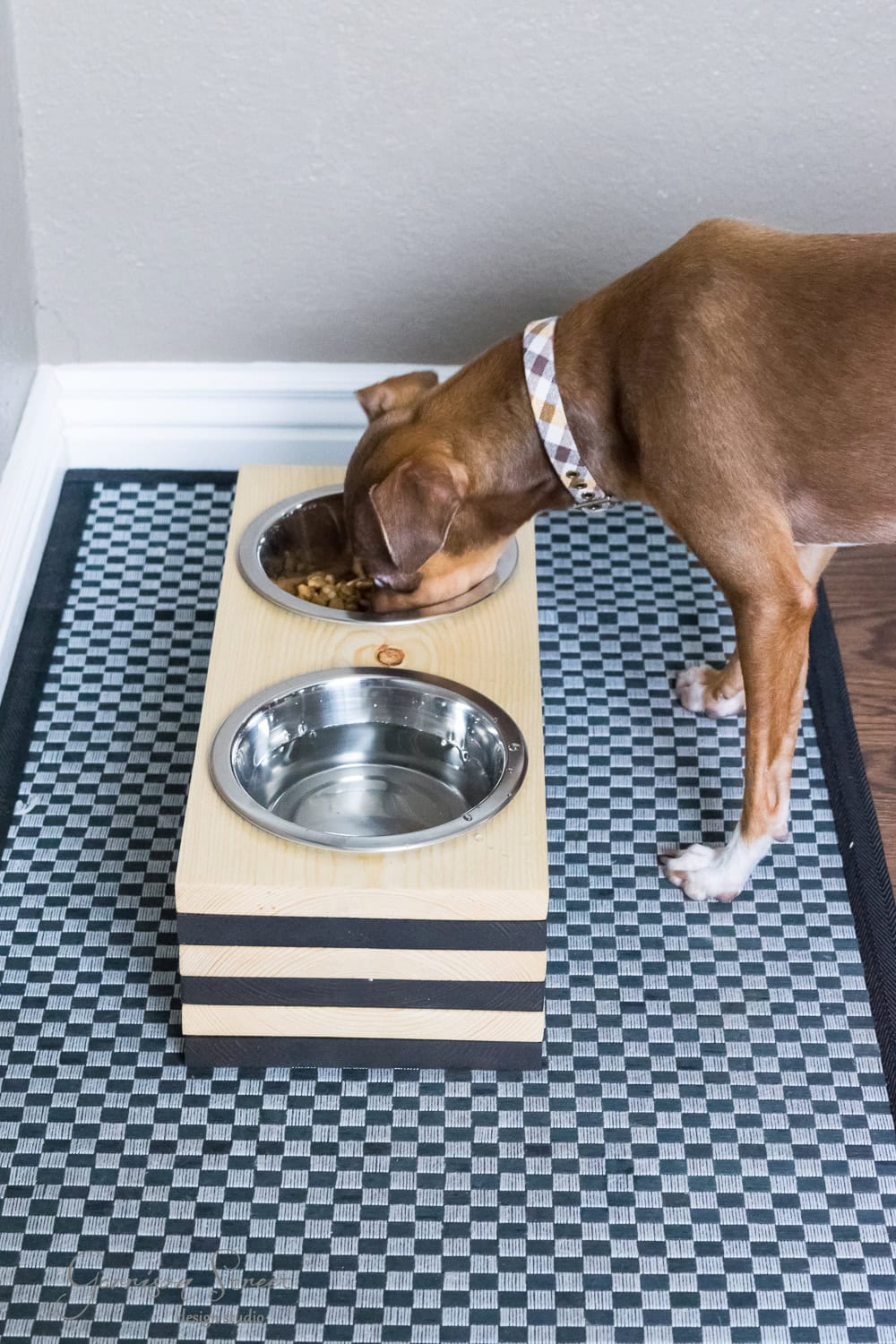 A brown dog with a patterned collar eagerly eats from a raised wooden stand holding two metal bowls—one filled with food and the other with water—resting on a checkered mat on the floor.