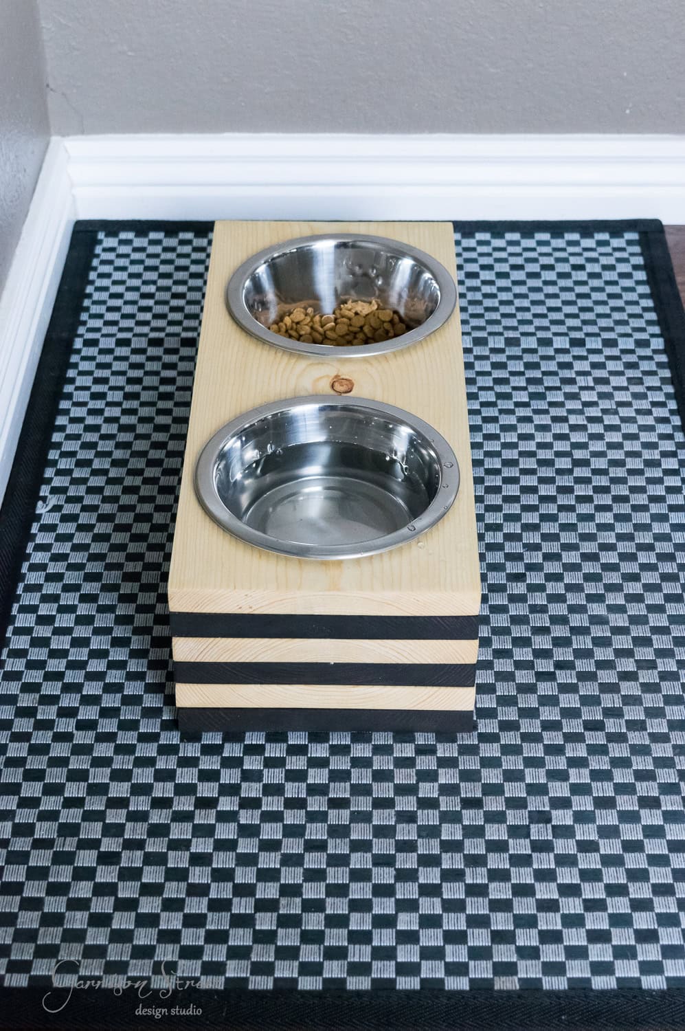 A raised wooden pet feeding station holds two stainless steel bowls on a black and white checkered mat. One bowl is filled with kibble, while the other contains water. Designed for dogs, it features a simple, modern look with multiple layers.