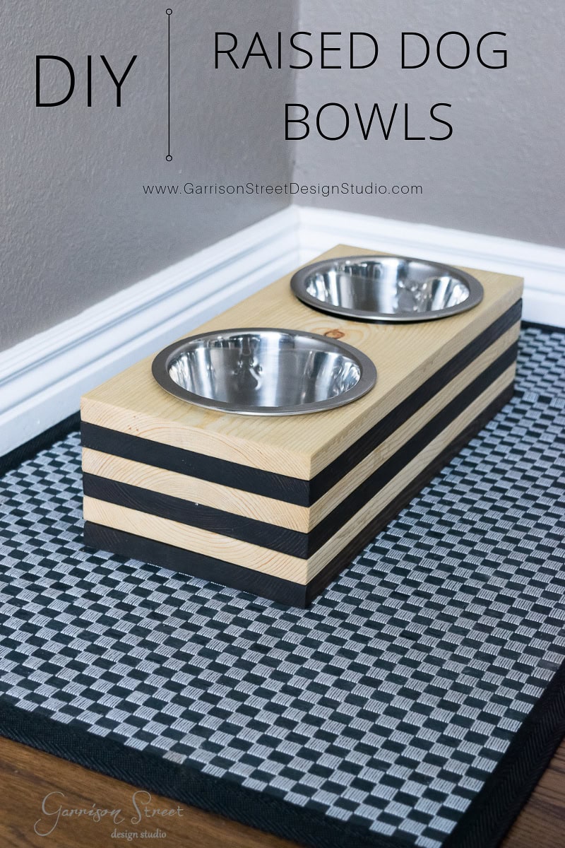 DIY raised dog bowls crafted from stacked wooden blocks featuring two metal bowls inset on top. Perfectly positioned on a black and white woven mat, these elevated bowls add style and comfort for your furry friend. Text reads "DIY Raised Dog Bowls.