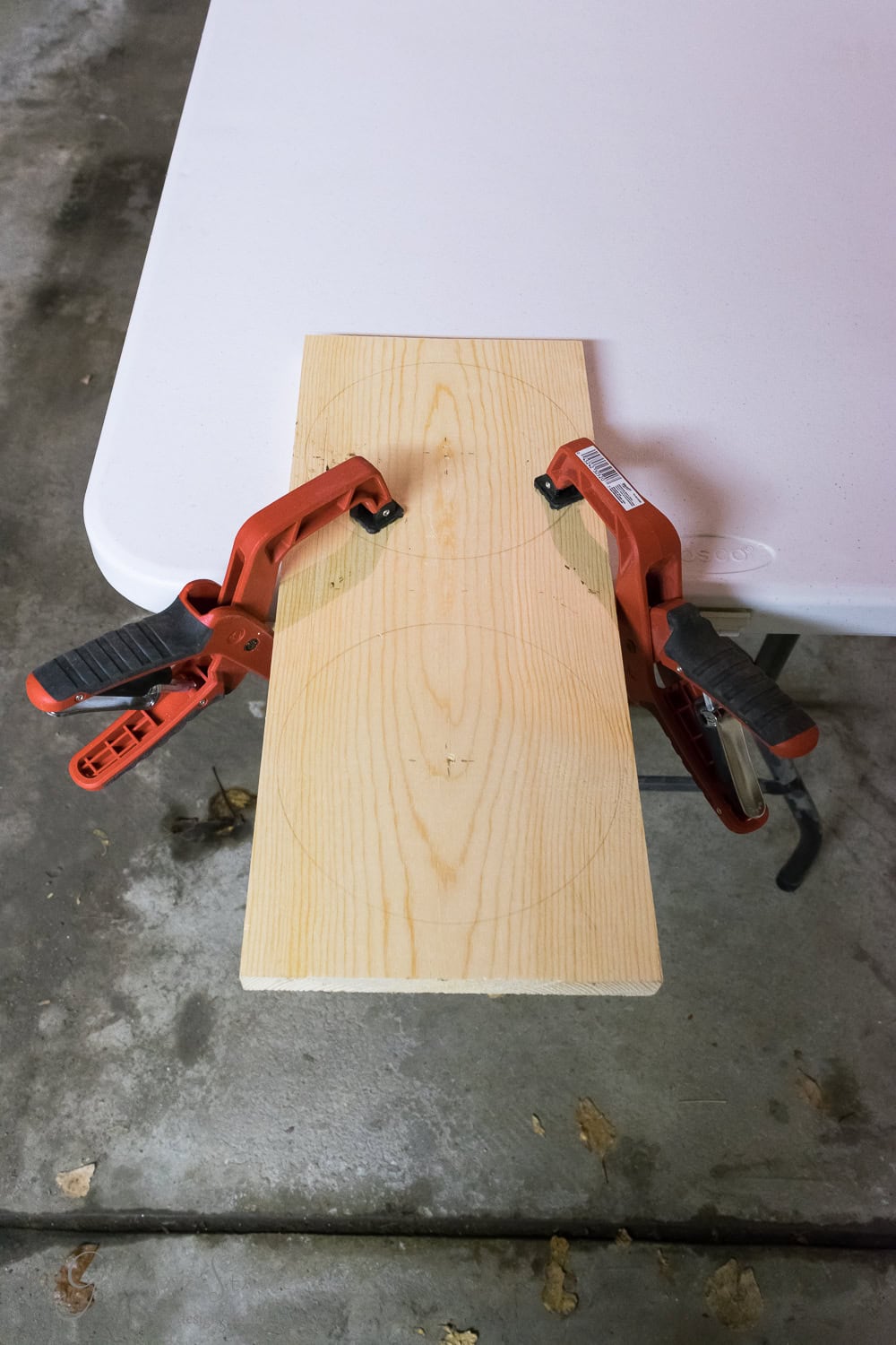 A wooden board with pencil marks, clamped to a white table using two red clamps, rests beside a raised bowl. The scene unfolds in a garage or workshop with a concrete floor and scattered leaves, hinting at the resident dog's playful presence.