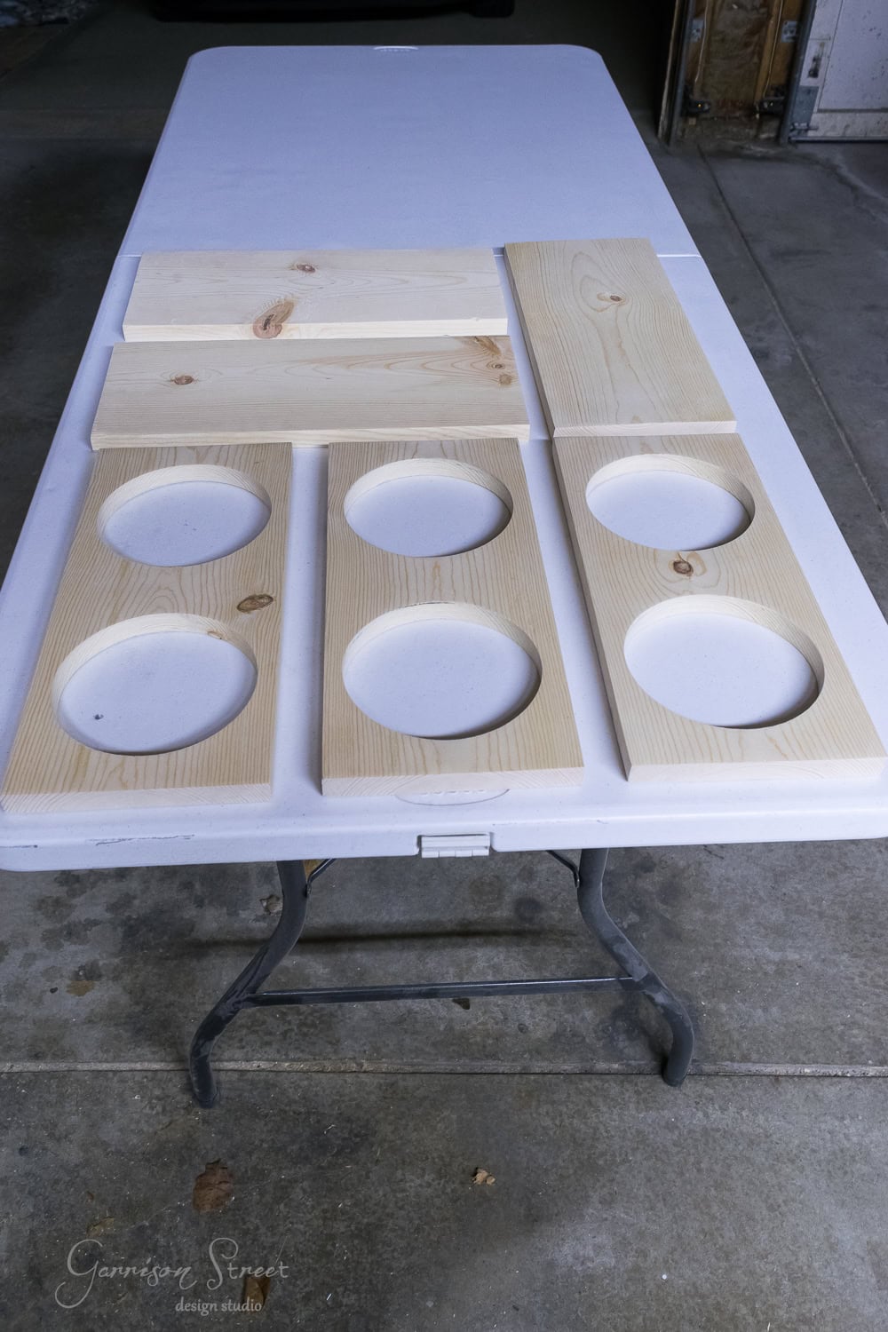 Wooden planks with circular cutouts are arranged on a white folding table in a garage. Some planks remain uncut, while others feature multiple indentations.