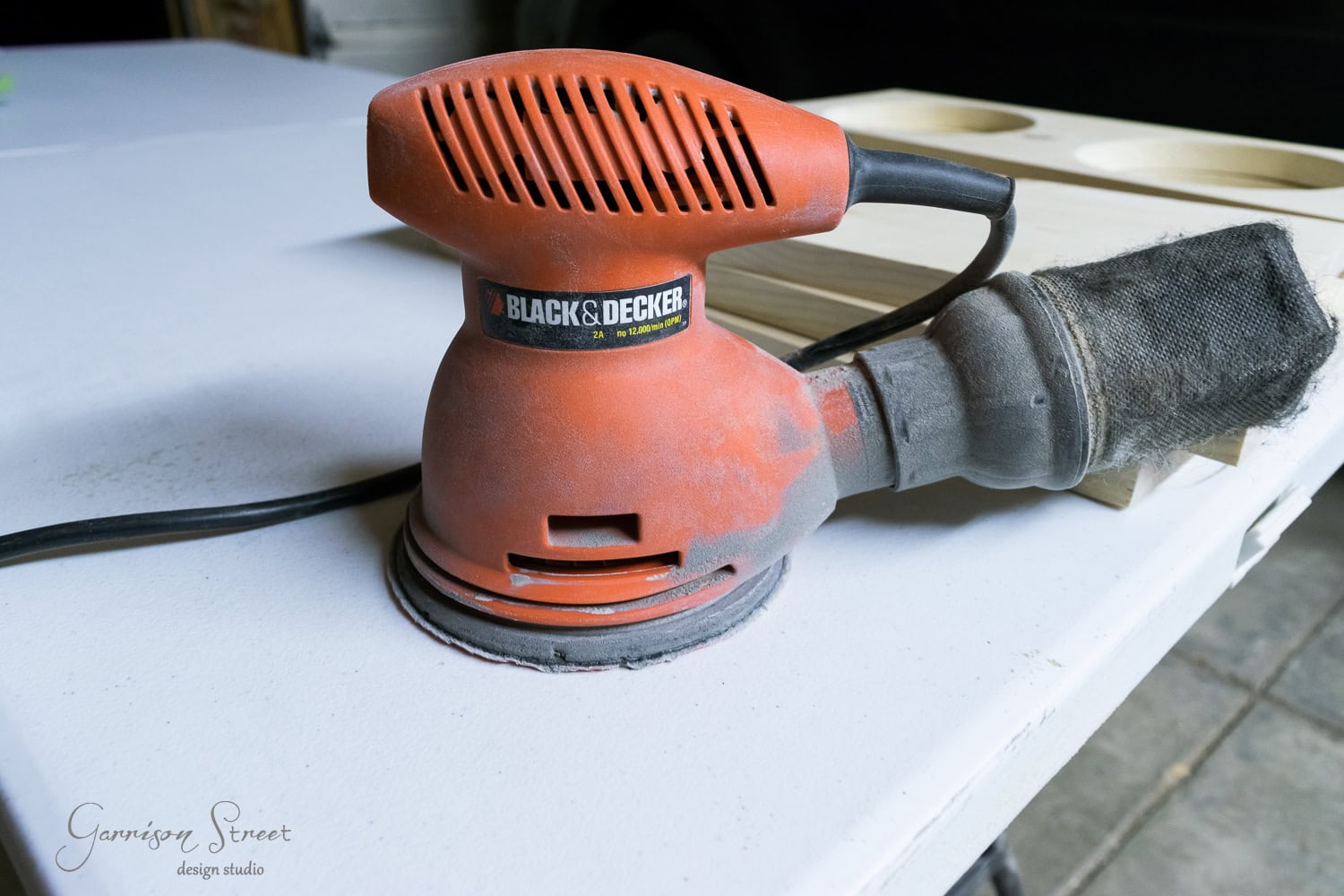 An orange Black & Decker palm sander rests on a white table, poised next to a bowl of sandpaper sheets. It's connected to a dust collection bag, with raised wooden boards in the background awaiting transformation.