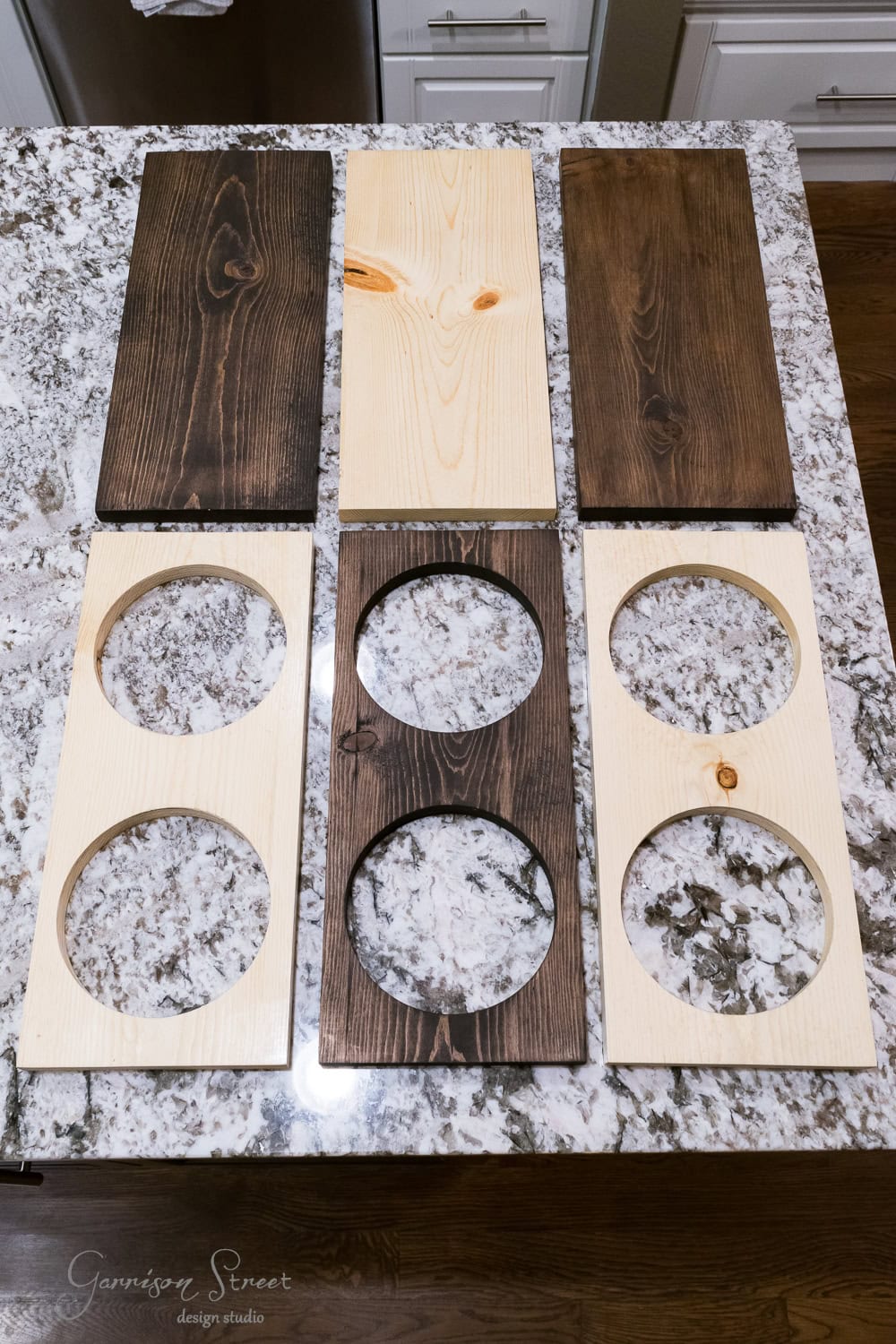 Two rows of wooden boards with circular cutouts are laid out on a speckled countertop. Some boards are stained dark brown, while others boast a natural light wood finish.