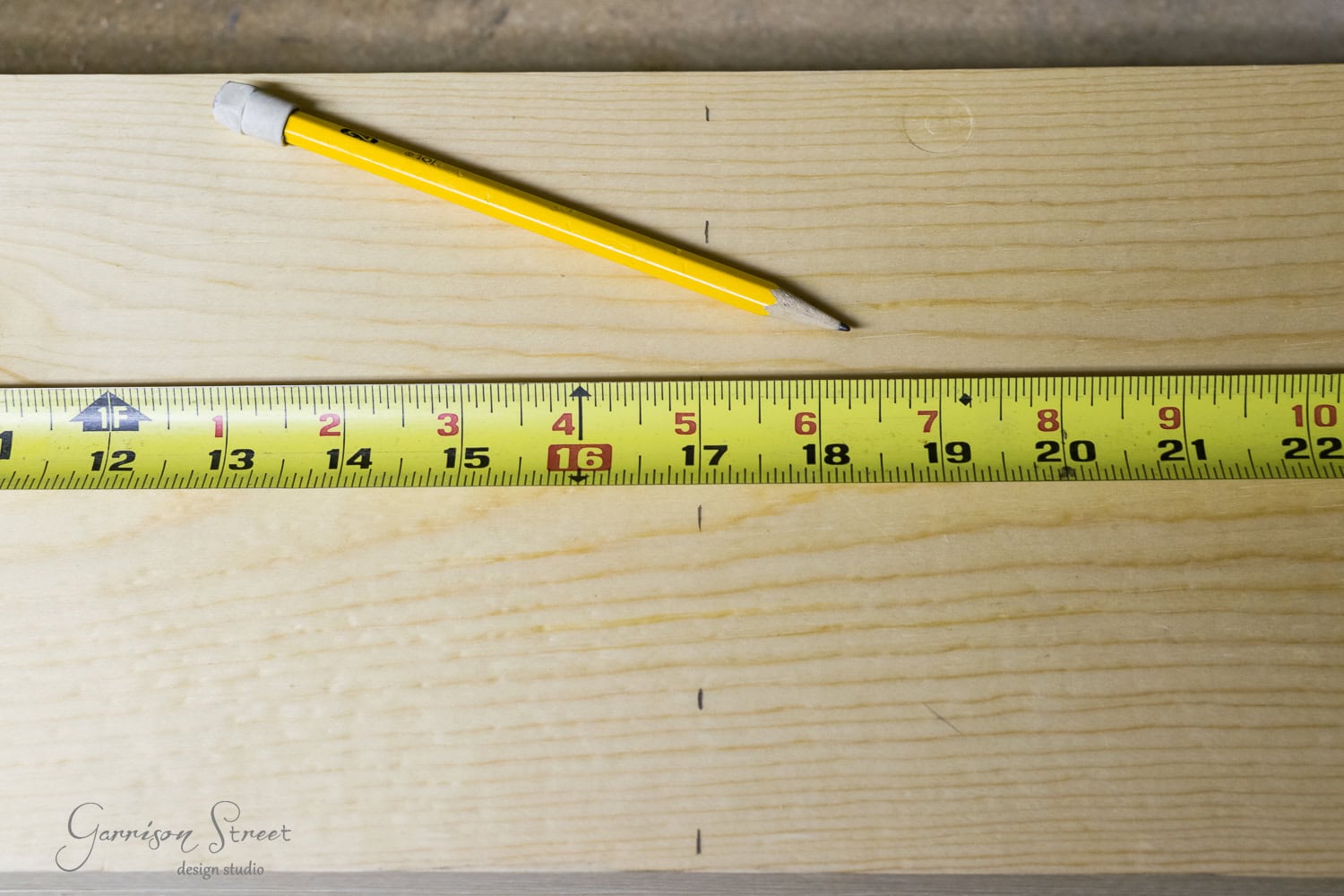 A yellow pencil and a yellow measuring tape are placed on a light wooden plank. The raised measuring tape marks, clearly visible, extend across the plank, indicating measurements in inches.