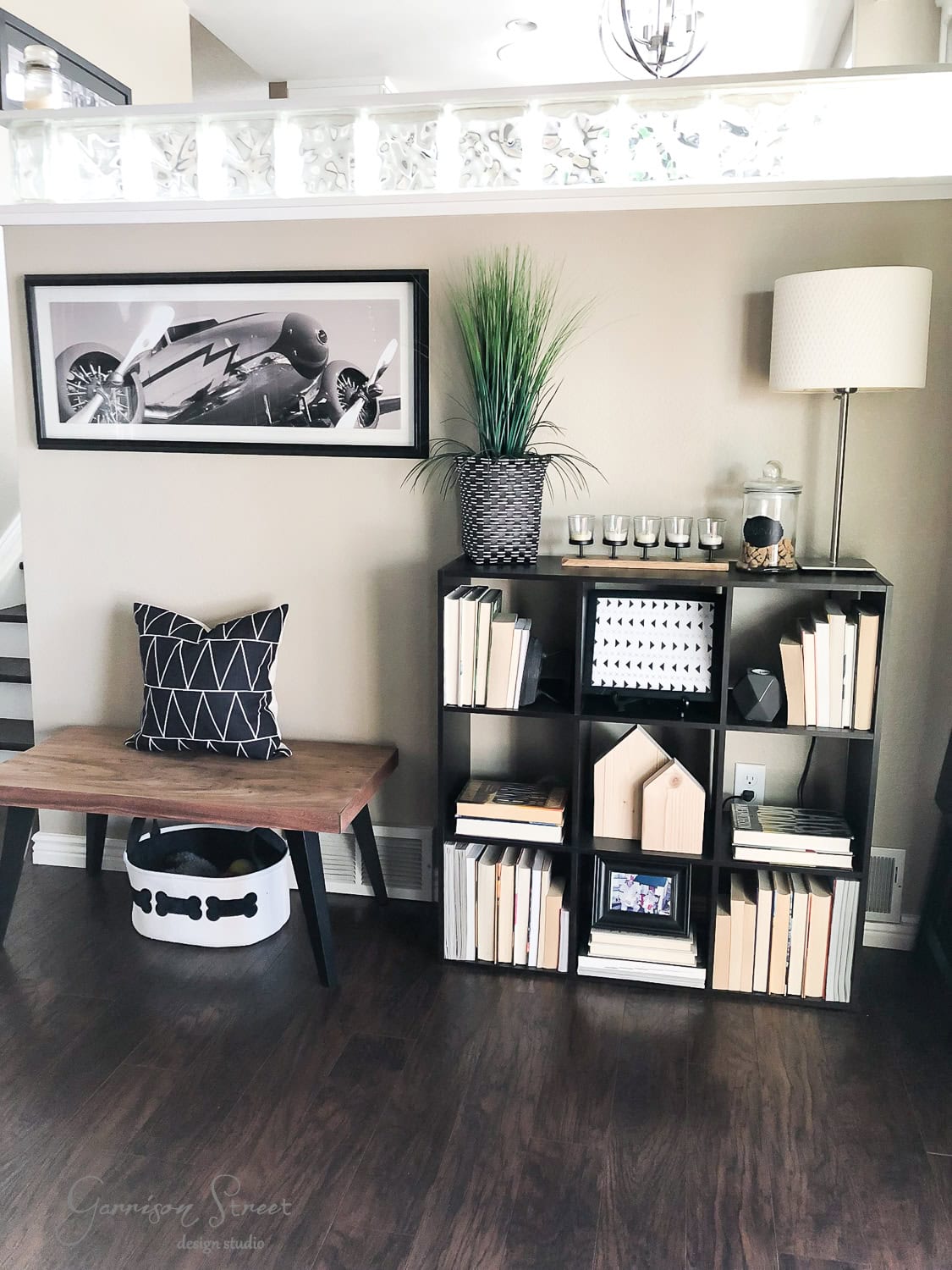 A modern living room with a dark wood floor features a bench adorned with a mud cloth patterned pillow, a cube shelf filled with books and decor, a tall plant, a lamp, and a black-and-white photo of a racing car on the wall.