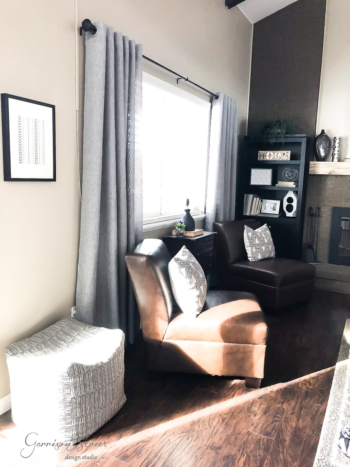 A cozy living room featuring two brown leather chairs and a mud cloth-patterned ottoman on a wooden floor. A window with gray curtains lets in natural light. A black bookshelf with decor items and a fireplace are in the background.