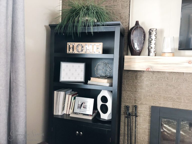 A black bookshelf stands in a cozy living room corner next to a stone fireplace. It displays books, decorative items, and a "HOME" sign. A plant sits atop the shelf, and curtains partially cover a window on the left.