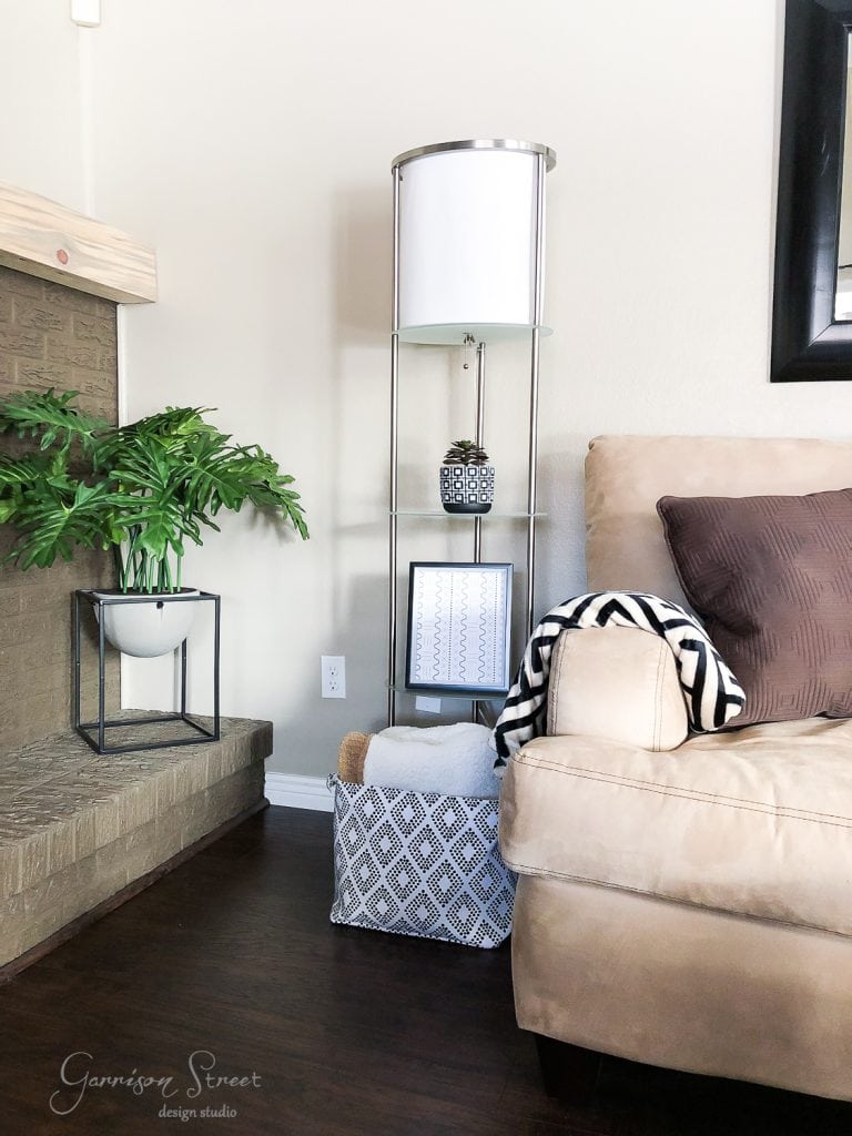 Free Printables A cozy living room corner featuring a beige armchair with a brown pillow, a chevron-patterned blanket, and a tall metal floor lamp. A decorative plant sits on the fireplace ledge while a basket adorned with mud cloth design rests on the floor, adding an earthy touch.