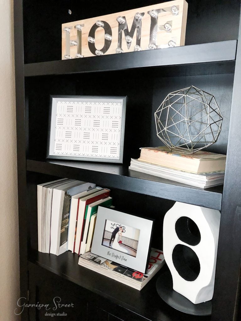 Free Printables A black shelf with a "HOME" marquee sign, geometric decor, framed art, and stacked books creates a neat and modern arrangement. A white sculpture sits on the bottom shelf beside free printables that add an artistic flair.