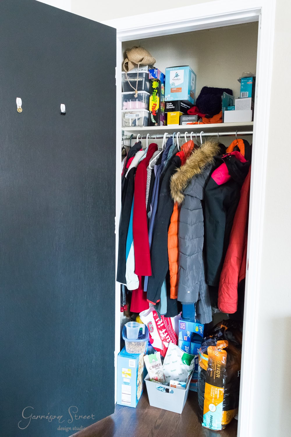 A closet with an open door reveals assorted coats hanging. The floor is cluttered with boxes, cleaning supplies, dog food, and household items. Shelves above hold storage bins and packaged goods—ideal for anyone seeking IKEA-inspired organization solutions.