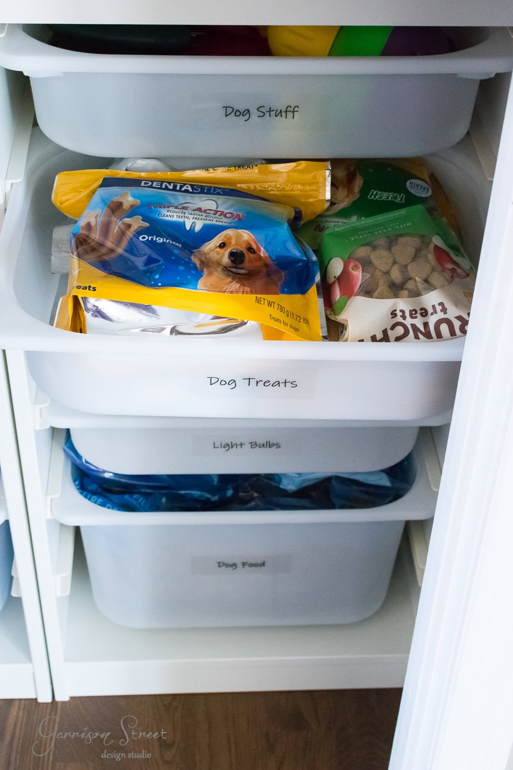 Open white cabinet with Trofast bins for seamless organization. Top bin: "Dog Stuff" with toys. Middle bin: "Dog Treats" containing a Dentastix bag and dog biscuits. Third bin: "Light Bulbs." Bottom bin: "Dog Food." Labels crafted in vinyl lettering bring order to your Ikea storage solution.