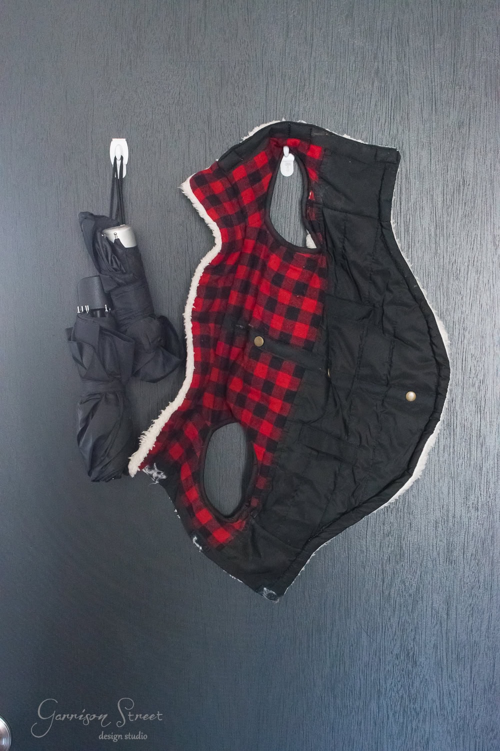 A black and red plaid dog coat hangs on a dark wood door, perfectly organized next to a folded black umbrella with a white handle, embodying the essence of an orderly closet.
