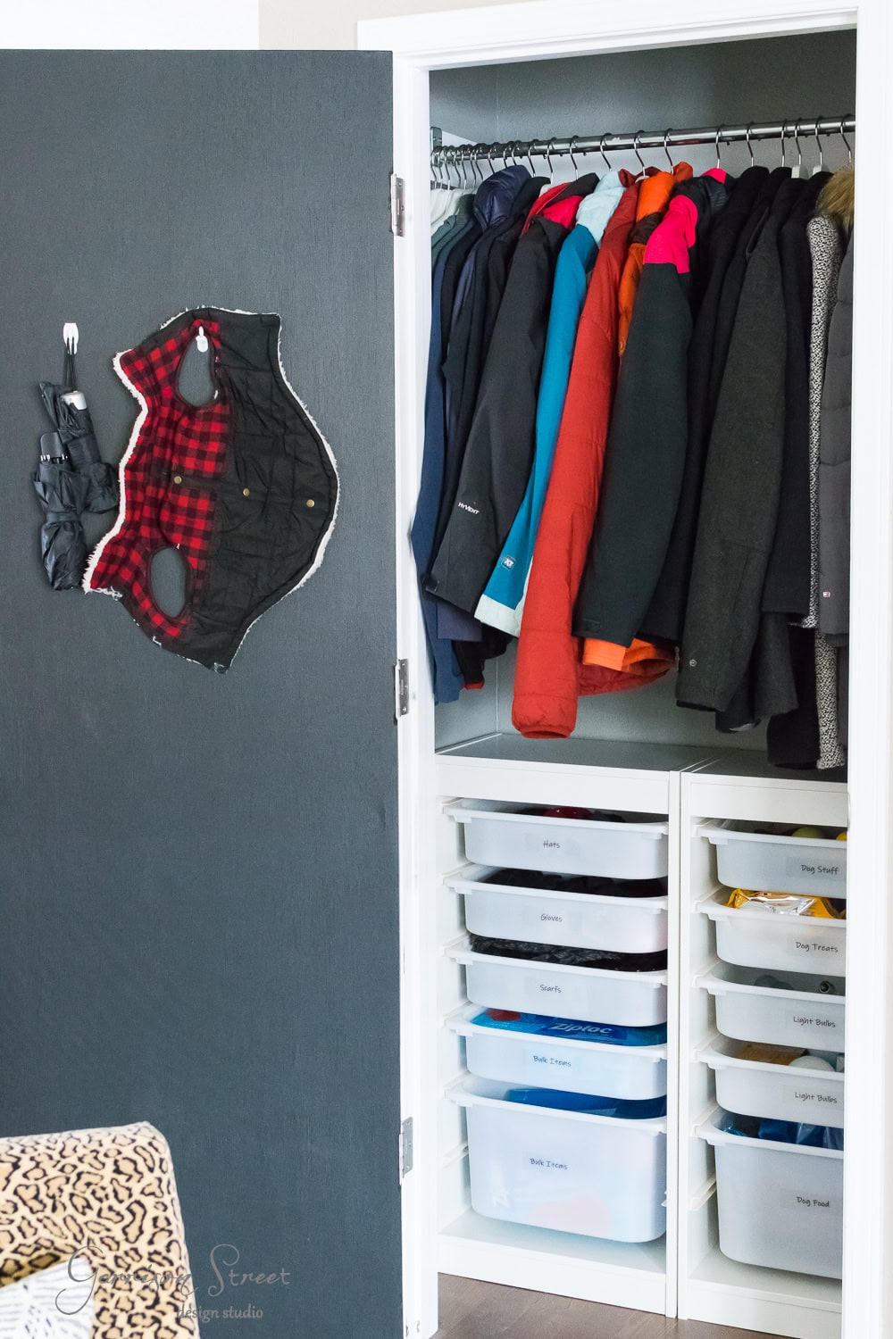 A neatly organized closet features winter coats hanging on a rod, with labeled IKEA Trofast drawers below for kids' clothes. A plaid face mask is casually hung on the inside of the door, highlighting efficient organization.