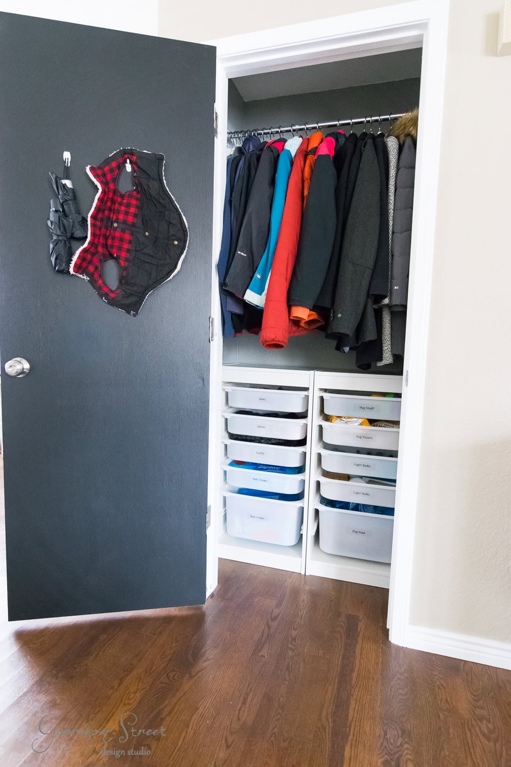 The small closet, featuring meticulous organization, displays jackets hanging neatly on a rod. Below, TROFAST white storage bins are perfectly arranged. Mounted on the open door is a sleek black wall organizer. The floor is wooden, completing the efficient and stylish space.