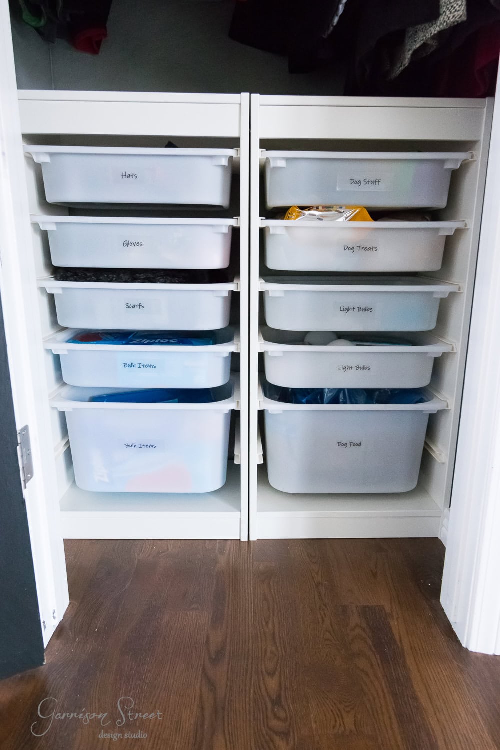 A neatly organized IKEA closet features two columns of white storage bins. Each bin is labeled with contents like hats, scarves, dog treats, and light bulbs. The closet is beautifully set against a dark hardwood floor, showcasing efficient organization.